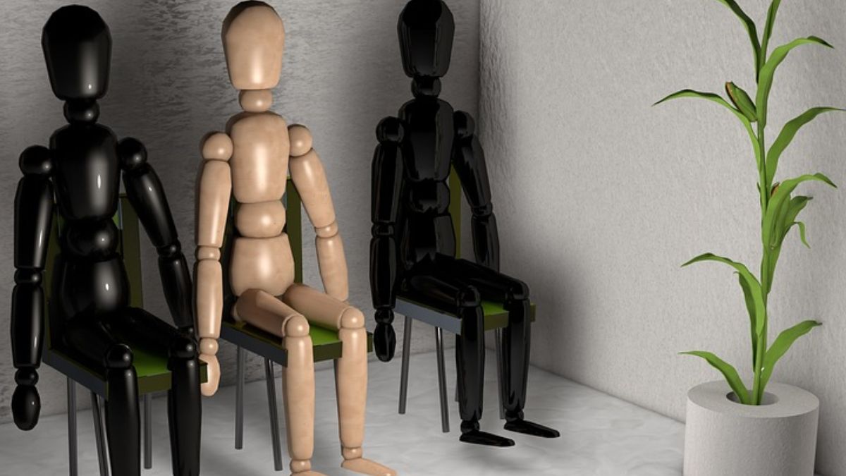 Three wooden mannequins posed on chairs in a waiting room, one light fawn-colored and two black, with a green potted plant nearby.