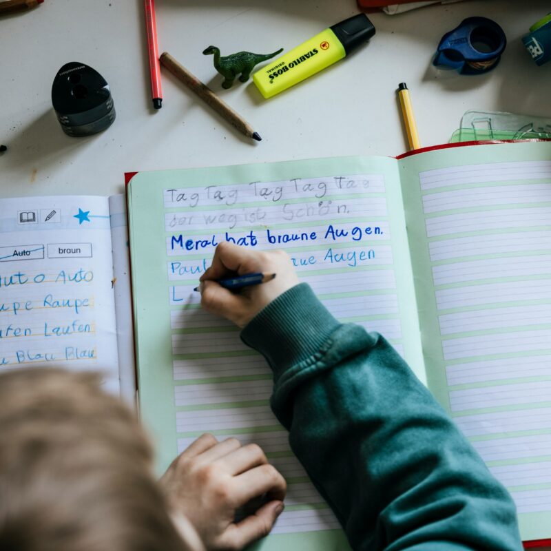 A child independently learning to write in cursive, using colored pencils on a notebook during an afternoon class.