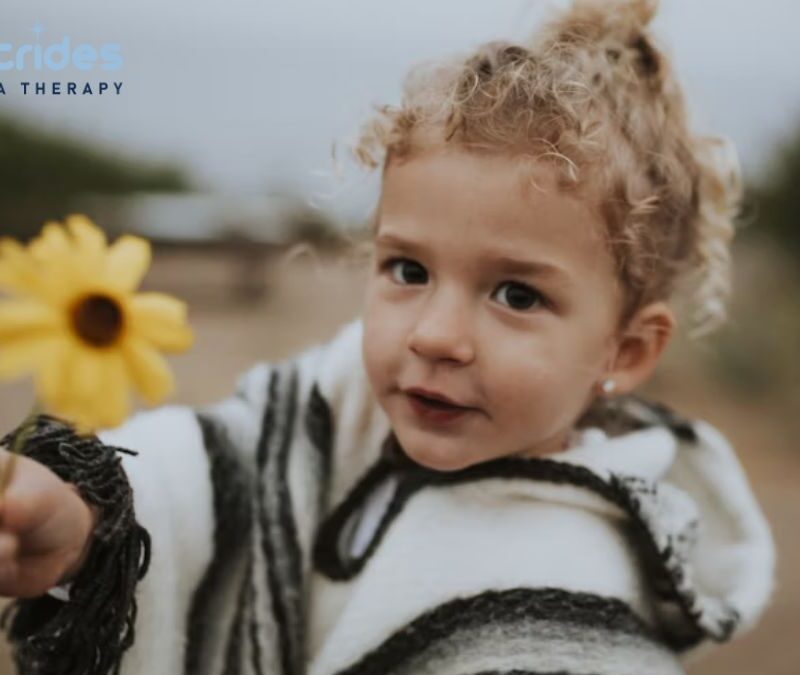 A young blonde girl with autism holds out a yellow daisy flower in the middle of a field under an overcast sky.