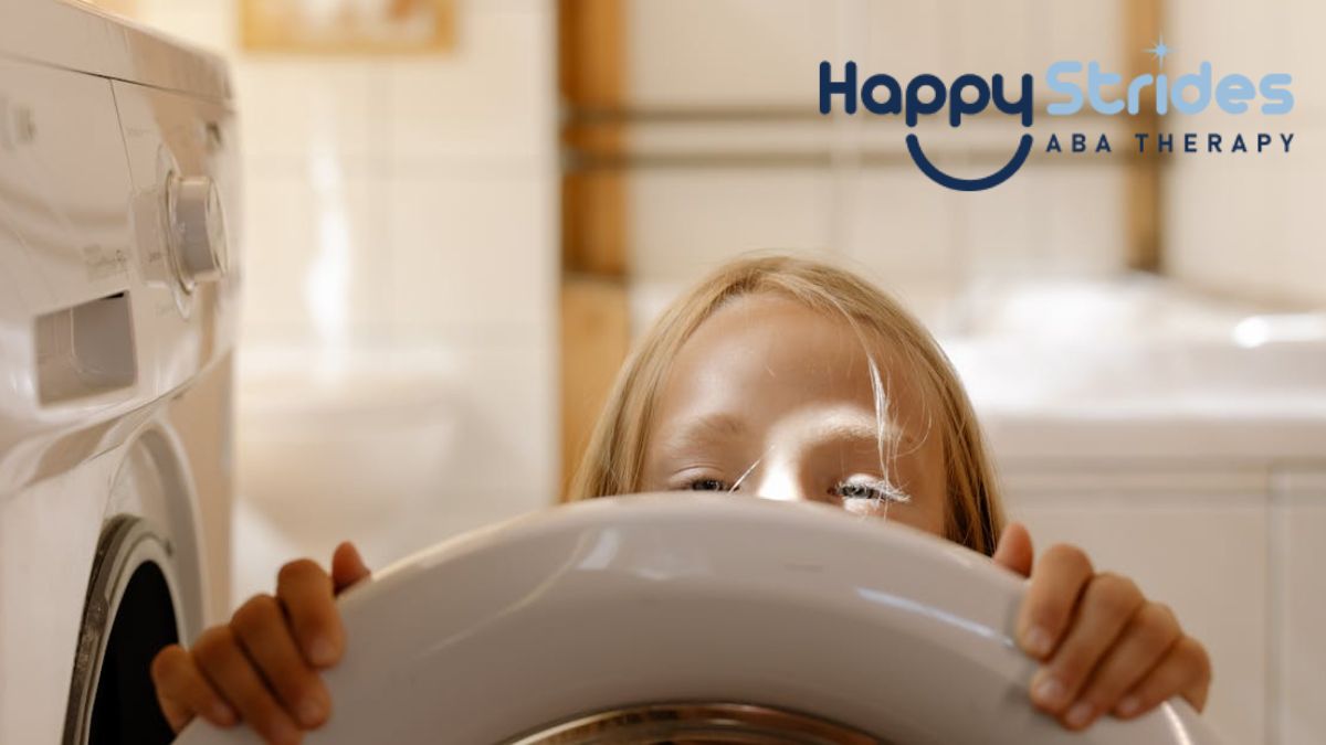 A young girl with autism peers through the door of a washing machine, showing her effort to help with household chores.