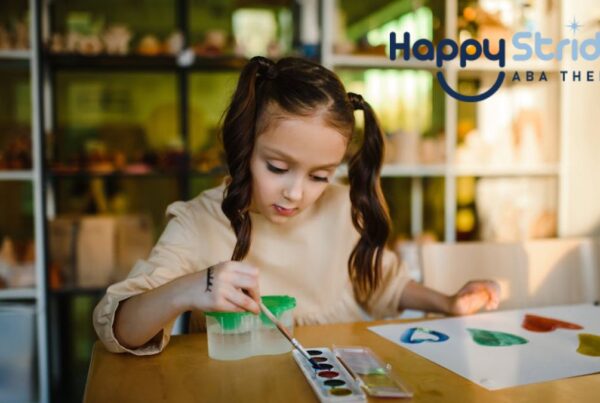 A young girl with autism sits at a table in a brightly lit room, fully focused on her painting, appearing capable and confident.