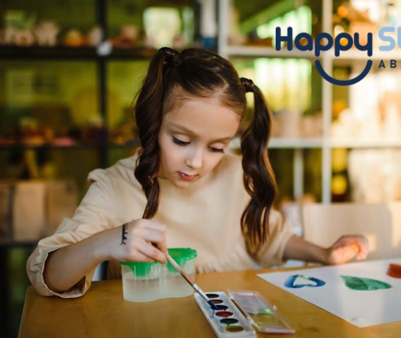A young girl with autism sits at a table in a brightly lit room, fully focused on her painting, appearing capable and confident.