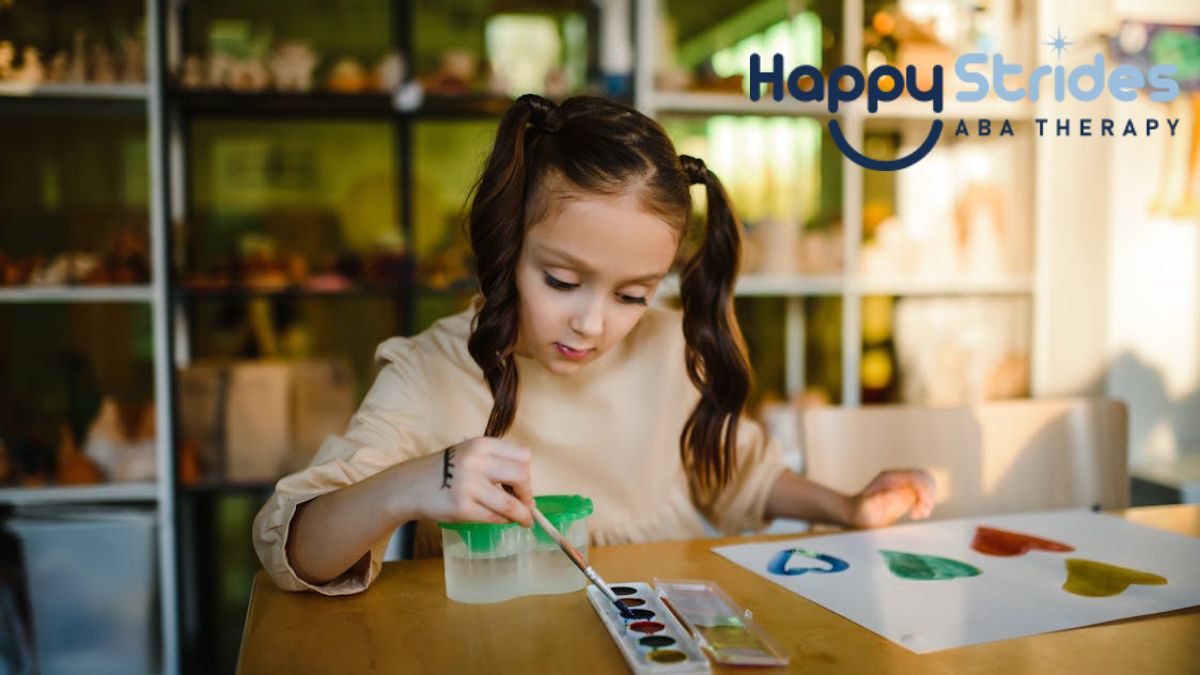A young girl with autism sits at a table in a brightly lit room, fully focused on her painting, appearing capable and confident.