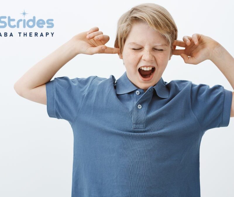 Hypersensitivity in autism, a boy in a blue shirt covers his ears while standing in a white room.
