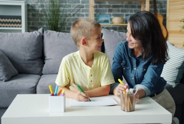 A parent and young boy engage in at-home autism therapy on the living room floor, with toys covering the coffee table in front of them.