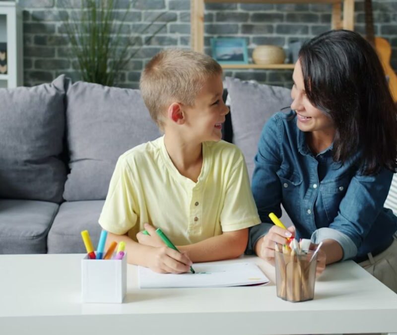A parent and young boy engage in at-home autism therapy on the living room floor, with toys covering the coffee table in front of them.
