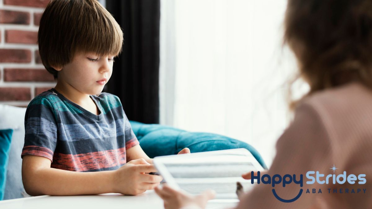 A young boy with nonverbal autism wearing a striped shirt looks down at his task while receiving therapy from an instructor whose back is to the camera.