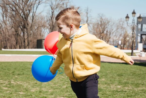 A young boy with autism spins in the middle of a grassy lawn, showing hyposensitivity behavior, while colorful balloons float in the background.