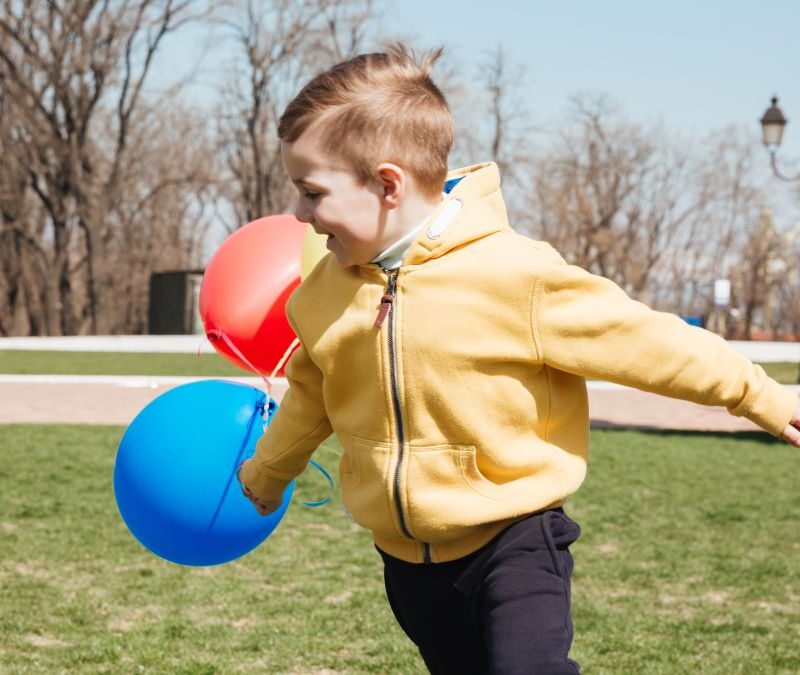 A young boy with autism spins in the middle of a grassy lawn, showing hyposensitivity behavior, while colorful balloons float in the background.