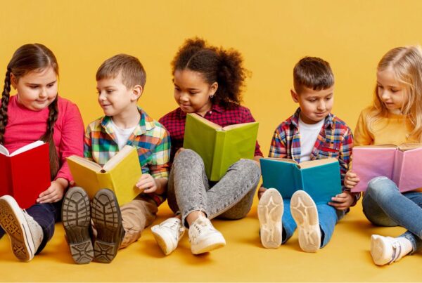 Children sitting in a line against a yellow wall, holding books and talking with each other, learning about ABA therapy.