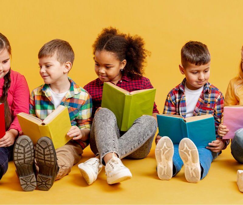 Children sitting in a line against a yellow wall, holding books and talking with each other, learning about ABA therapy.