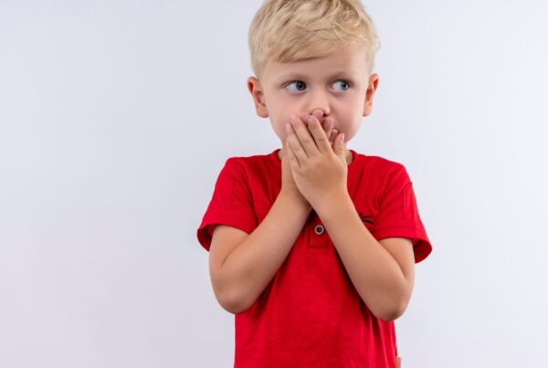 A young boy with nonverbal autism wearing a red shirt covers his mouth with both hands against a plain white background.