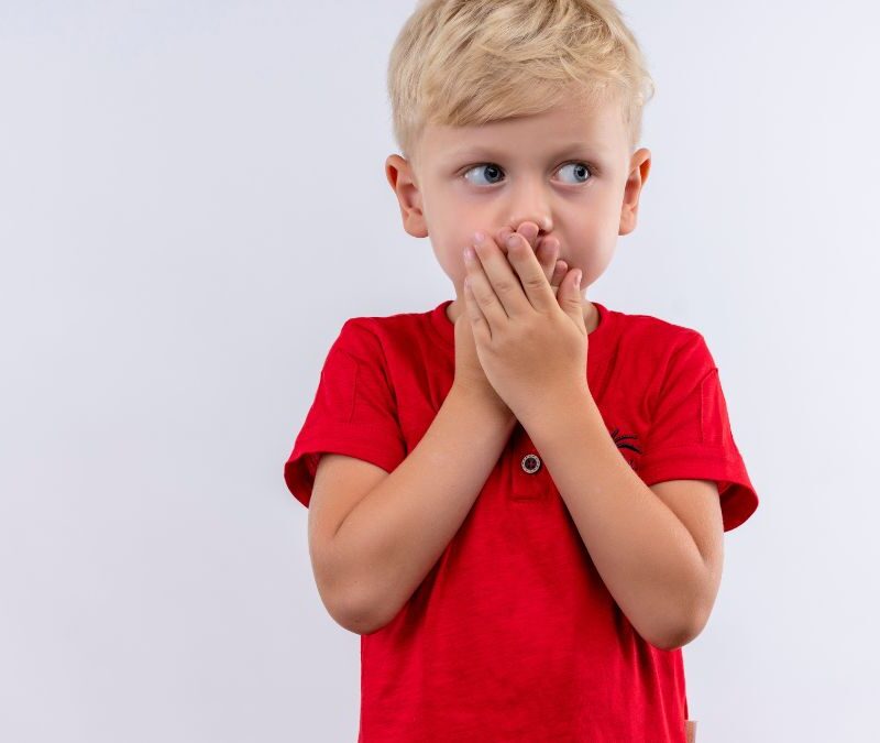 A young boy with nonverbal autism wearing a red shirt covers his mouth with both hands against a plain white background.