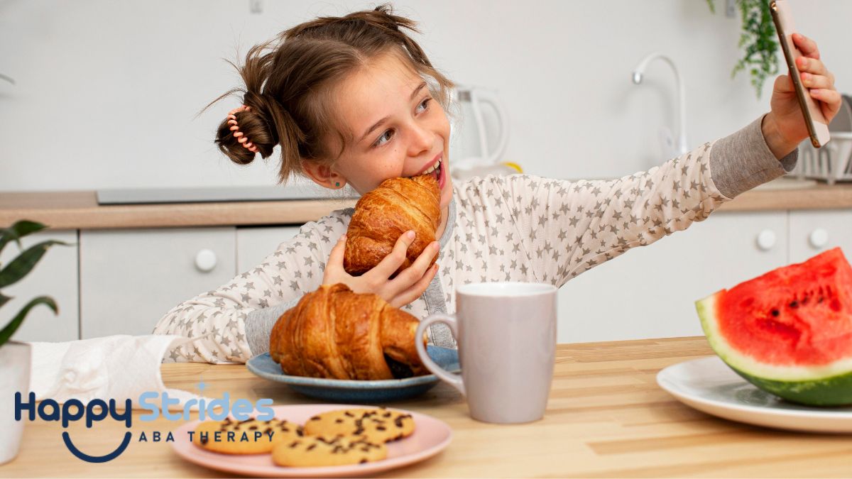 A young girl takes a bite of a croissant while holding up a smartphone to take a selfie, seated at a table filled with various mealtime foods during an ABA food therapy session.