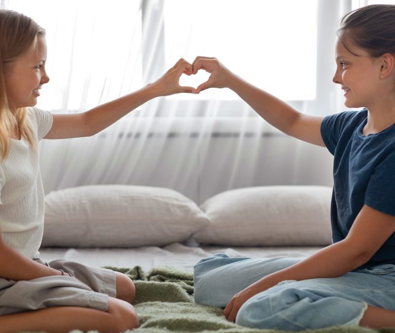 Two young girls sit on a bed, smiling as they form a heart shape with their fingers during an autism self-help activity.