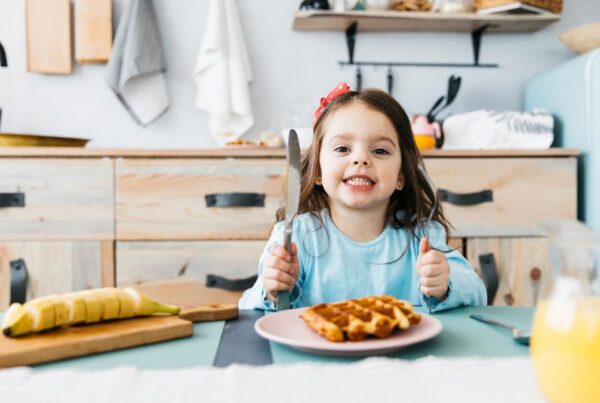 A child wearing a blue blouse smiles confidently at the camera while sitting at a table filled with breakfast foods during an ABA food therapy session.