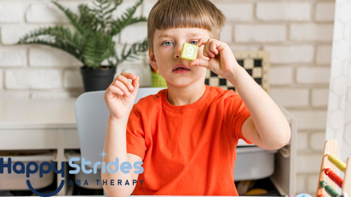 A boy with autism holds up a visual cue card near his eye, with a table in front of him strewn with other cards and pieces, practicing emotional awareness.