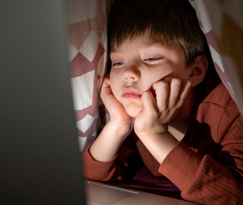 Boy staying up late while using a laptop, appearing tired in a dimly lit room.