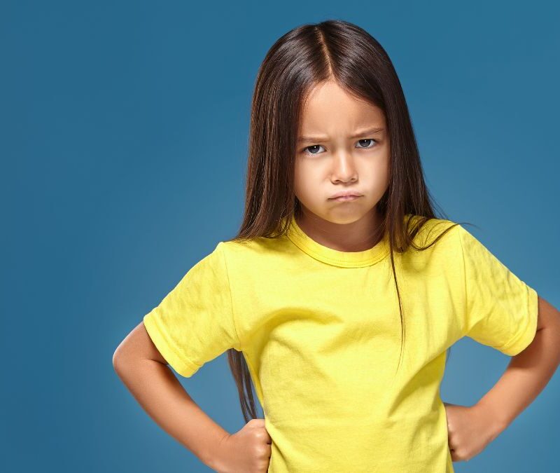 A young girl with autism wearing a yellow shirt frowns at the camera, showing signs of emotional awareness.